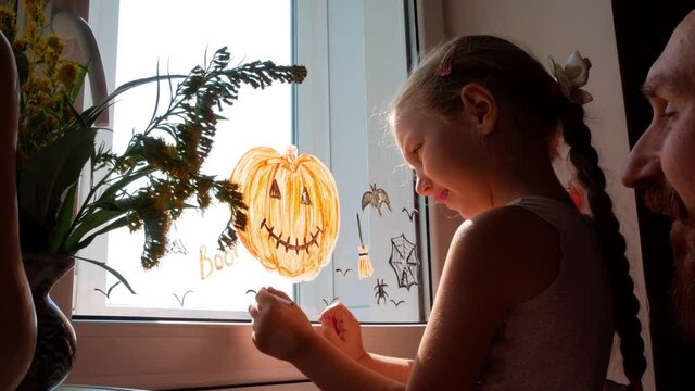 Child with father painting pumpkin on window preparing to Halloween Little girl with dad drawing decorating room paper bats celebration autumn holiday home.Creative family leisure lockdown new reality