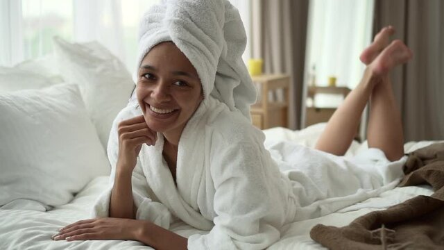 Beauty and relaxing in hotel room. Spbd Smiling African-American woman wearing terry bathrobe with turban on head lies on large bed in light room