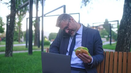 Busy businessman eating sandwich while working on laptop in park, unhealthy food - Powered by Adobe