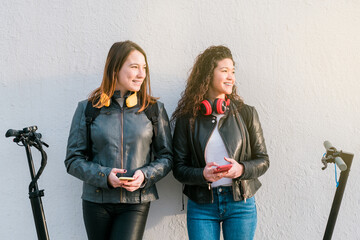 Two multiethnic female friends on electric scooters using smartphone outdoors. Two women standing over a white wall with headphones. Urban lifestyle and ecological transport concepts - Powered by Adobe