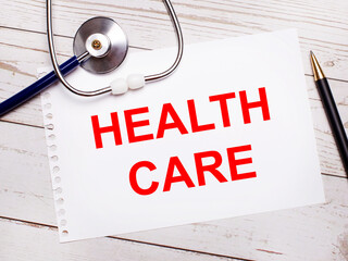 On a light wooden table there is a stethoscope, a pen and a sheet of paper with the text HEALTH CARE. Medical concept