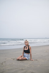 Pretty fit caucasian young woman with blond hair, wearing denim shorts, short top and stylish necklace. Sitting on the beach. Looks at camera. 