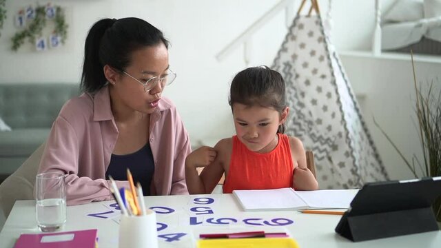 Home schooling for children. Spbd Thoughtful little Korean girl looks for card with digit solving sums with mother near tablet in light room against toy wigwam