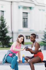 Fototapeta na wymiar Cheerful smiling friends in sportswear sitting on a bench in the city discussing while using smartphone looking to screen. Multiethnic women having a fitness workout break.