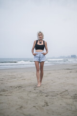 Portrait of pretty caucasian young woman with blond hair, wearing denim shorts, short top and stylish necklace. Walking on the beach. Looks at camera