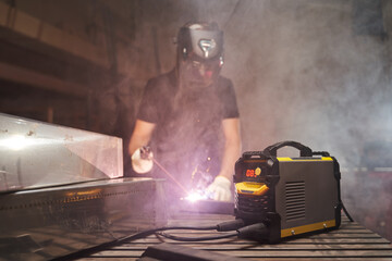 a welder in a helmet and gloves in smoke works with an inverter welding machine