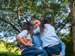Fototapeta premium Two sibling little girls with mom laughing and hugging each other on warm and sunny summer day in the garden. Young girls with her mother spending day in park.