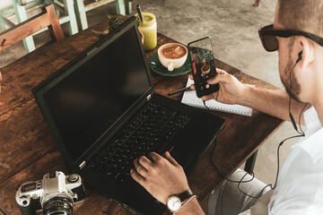 Człowiek pracujący przy laptopie z smartfonem, piszący notatki w zeszycie przy stole. - obrazy, fototapety, plakaty