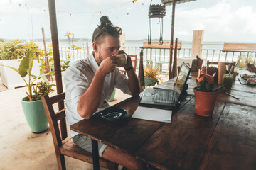 Mężczyzna, cyfrowy nomada pracujący z laptopem i telefonem przy stoliku w kawiarni, praca zdalna podczas podróży.
