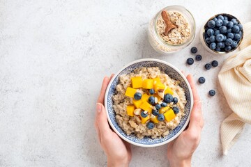Oatmeal bowl with fruits and berries in a young woman's hands. Cooked oats porridge with mango and...