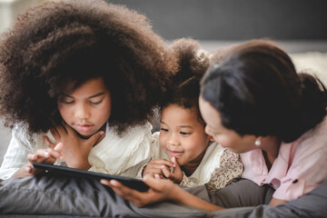Cheerful young mother sitting on couch at home with two beautiful daughter having curly hair and laughing while using digital tablet with children