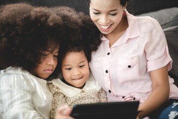 Cheerful young mother sitting on couch at home with two beautiful daughter having curly hair and laughing while using digital tablet with children