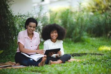 Cheerful young beautiful mother sitting with son with curly hair in park and helping him while using digital tablet while studying