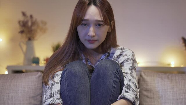 close up girl sitting with arms around knees and a tv controller is frightened and peeping through fingers while seeing a horror film alone at home.