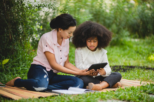 Cheerful young beautiful mother sitting with son with curly hair in park and helping him while using digital tablet while studying