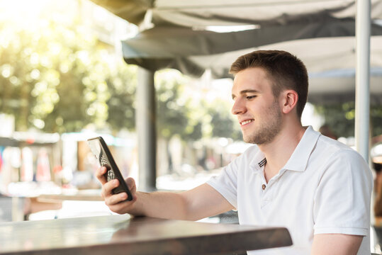 Young Man Looking At His Mobile Phone Cell Phone On The Terrace Of A Bar