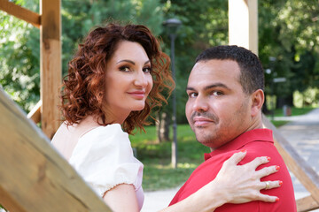 Loving couple on park bench turned around and looked at the camera. Man in red T-shirt and a curly woman in a white dress on a date. Woman put her hand on her husband's shoulder. Romantic summer walk