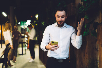 Excited young bearded man reading text message on smartphone in contemporary cafe