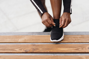 Afro-American woman hand tying sport black shoe lace while holding foot on a wood bench outdoors. Multiethnic women having a fitness workout break.
