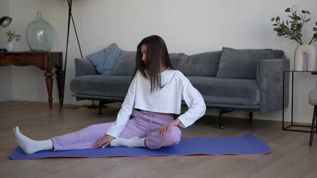 Beautiful slim young woman doing sports on a blue sports mat, home interior with grey sofa and decoration. Woman in sport clothes stretch body on a mat, gimbal shot