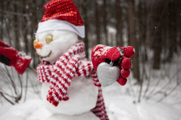 Obraz na płótnie Canvas snowman in a Santa hat, a striped scarf and red gloves is holding a souvenir silver heart. Snowman out of focus. Winter season in a snowy forest. Christmas holidays with love