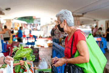 older woman with gray hair and medical protection mask buying vegetables in a traditional market.