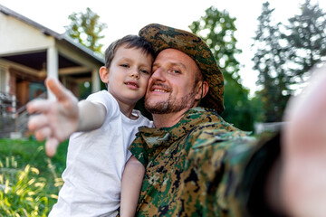 Man in military uniform making a selfie with smartphone with his lovely son. Soldier taking selfie with his little boy. Smiling father in military uniform and son having fun taking a selfie in nature.