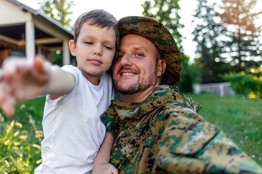 Young soldier father and his adorable little son taking selfie in the front of their house. A loving family having fun and creates memories by taking pictures with the smartphone, outdoor.