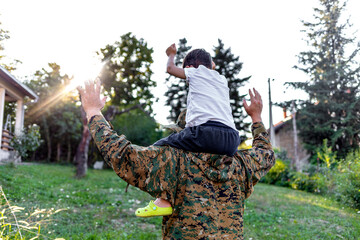 Cheerful dad is glad to be home from military assignment, he is carrying his son on his shoulders. Young military men holding his son on the shoulders with arms raised up in the air, looking away.