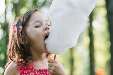 Little cute girl 3-4 eating cotton candy in sunny park among tall trees on green grass