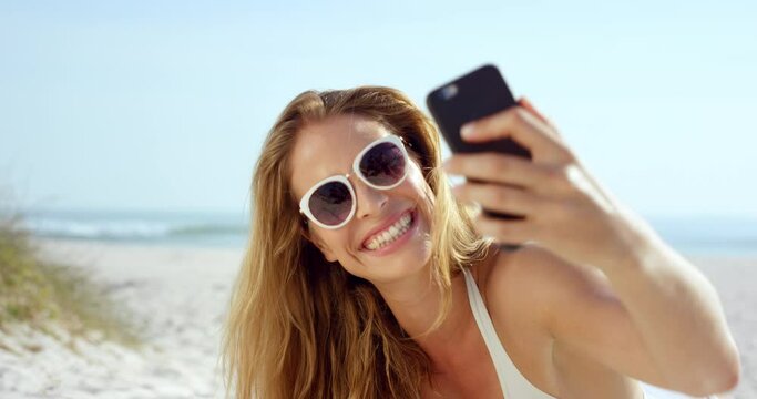 Woman talking selfie using phone sitting on beach wearing designer one piece swimsuit