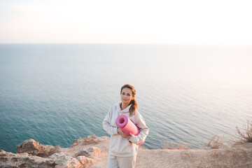 Smiling cheerful woman 25-26 year old hold yoga mat wear training hoodie posing over sea nature background. Healthy active lifestyle. Healthcare. Adult girl practice fitness outdoors. Happiness. 20s.