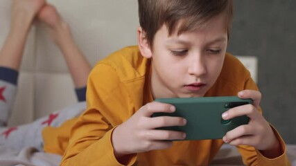 Cute little kid boy Laying on the bed focused on smartphone. Boy plays in video game on his smartphone. Child has fun playing videogame. Social and technology concept