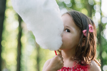 Little cute girl 3-4 eating cotton candy in sunny park, among tall trees on green grass