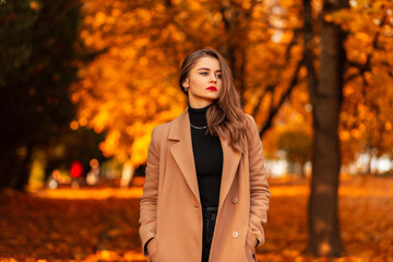 Pretty girl with red lips in a fashionable coat with a black vintage sweater walks in amazing autumn park with orange foliage at sunset