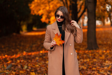 Stylish business woman model with vintage sunglasses in fashion beige coat with orange-red colored leaf walks in park with bright fall foliage