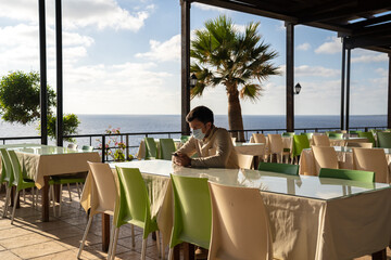Lonely sad masked man sits and uses smartphone himself in empty restaurant in cyprus at Agios Georgios. Visitor wearing mask waits order while sitting in deserted cafe, social distance and quarantine