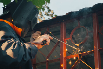 Close up of a welder hands welding steel structure metal part in home backyard. Craft man use electric arc argon wear work uniform,protective helmet on head without gloves while sparks fly at factory.