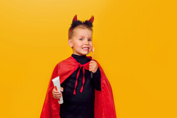 One little toddler boy in a carnival costume with toothpaste and an orange brush for Halloween is isolated on a yellow background. Medicine, dental hygiene, holidays concept.
