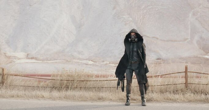 Post apocalyptic human, woman in grunge clothes hooded black mantle protective mask standing by hills in desert. Cyberpunk steampunk soldier with gun. Future female survivor. Post apocalyptic world