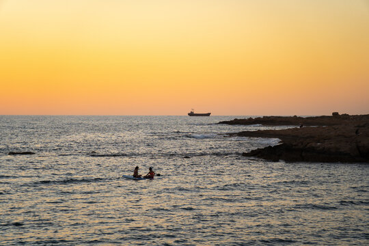 Two women sharing paddle board at sunset on calm waters in mediterranean sea in cyprus. Two girls on paddling on SUP. Silhuette in the distance. Water sport, sup surfing, surfboard or paddle boarder