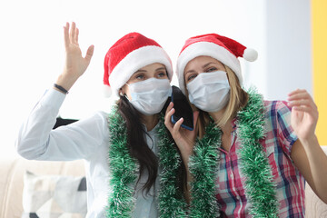 Women in red santa claus hats and protective medical masks talking on cell phone at home