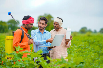 Young indian agronomist giving liquid fertilizer bottle to farmer and showing product information in laptop at green agriculture field. - obrazy, fototapety, plakaty