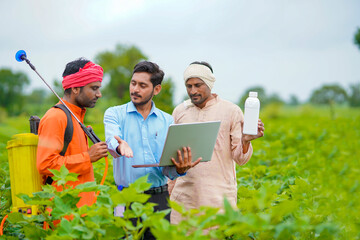 Young indian agronomist giving liquid fertilizer bottle to farmer and showing product information in laptop at green agriculture field.