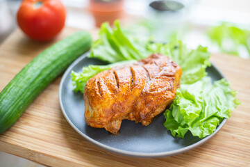 Grilled poultry fillet lies on a plate with lettuce. Nearby vegetables: cucumber and tomato.