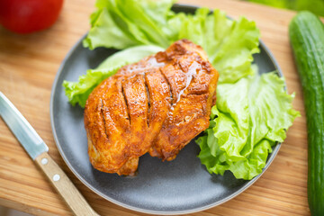 Grilled poultry fillet lies on a plate with lettuce. Nearby vegetables: cucumber and tomato.