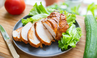 Grilled poultry fillets are sliced on a plate with lettuce. Nearby vegetables: cucumber and tomato.
