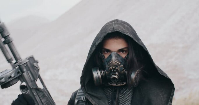 Portrait post apocalyptic human, woman in grunge clothes hooded black mantle protective gas mask respiratory standing by hills in desert. Cyberpunk soldier holding gun. Future female survivor