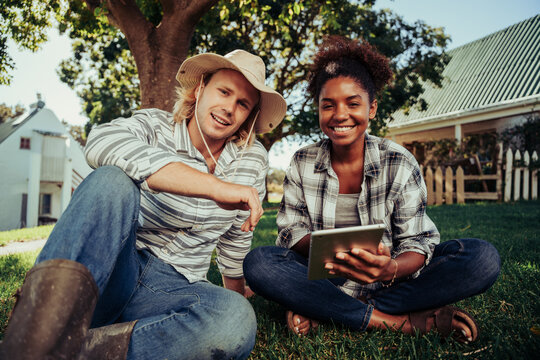 Mixed race couple smiling while enjoying time together sitting on grass scrolling through images on digital device 