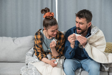 Young couple suffering from a common cold and flu sitting at home wrapped in blankets and wipe their noses with tissues while they have a strong headache, healthcare concept